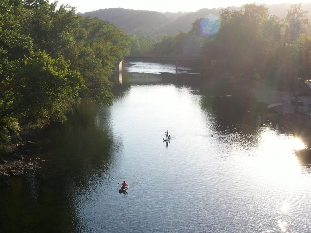Paddlers on the Greenbrier River near Lewisburg, West Virginia, gliding through calm, reflective water at sunset beneath a riverside bridge and tree-lined banks.