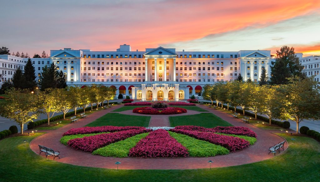 Historic Greenbrier Resort at sunset in Greenbrier Valley, West Virginia, showcasing its grand white facade, glowing entrance lights, and vibrant flower gardens.