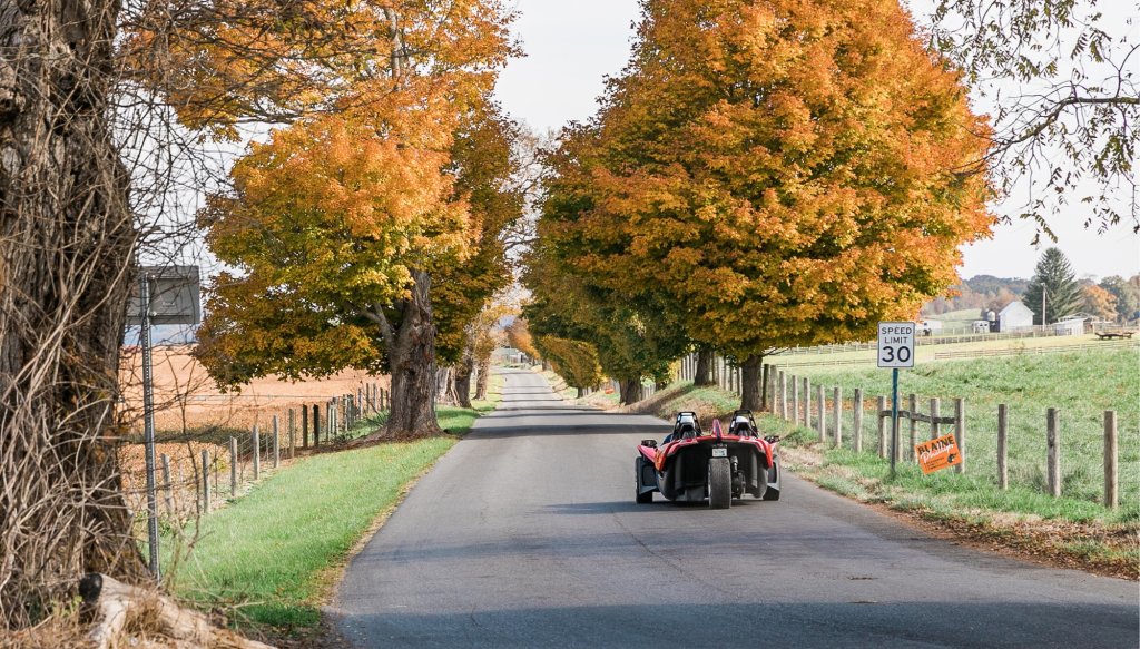 Scenic fall drive in Greenbrier Valley, WV, with a red roadster on a tree-lined country road surrounded by vibrant autumn colors.